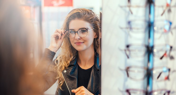 Smiling young woman trying on glasses on mirror in optician.