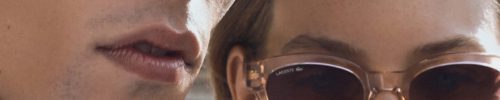 Close-up of a man and woman wearing Vu sunglasses outdoors, with buildings blurred in the background. Both have neutral expressions as sunlight highlights their faces, showcasing the chic style from le plus grand inventaire de montures à Montréal.
