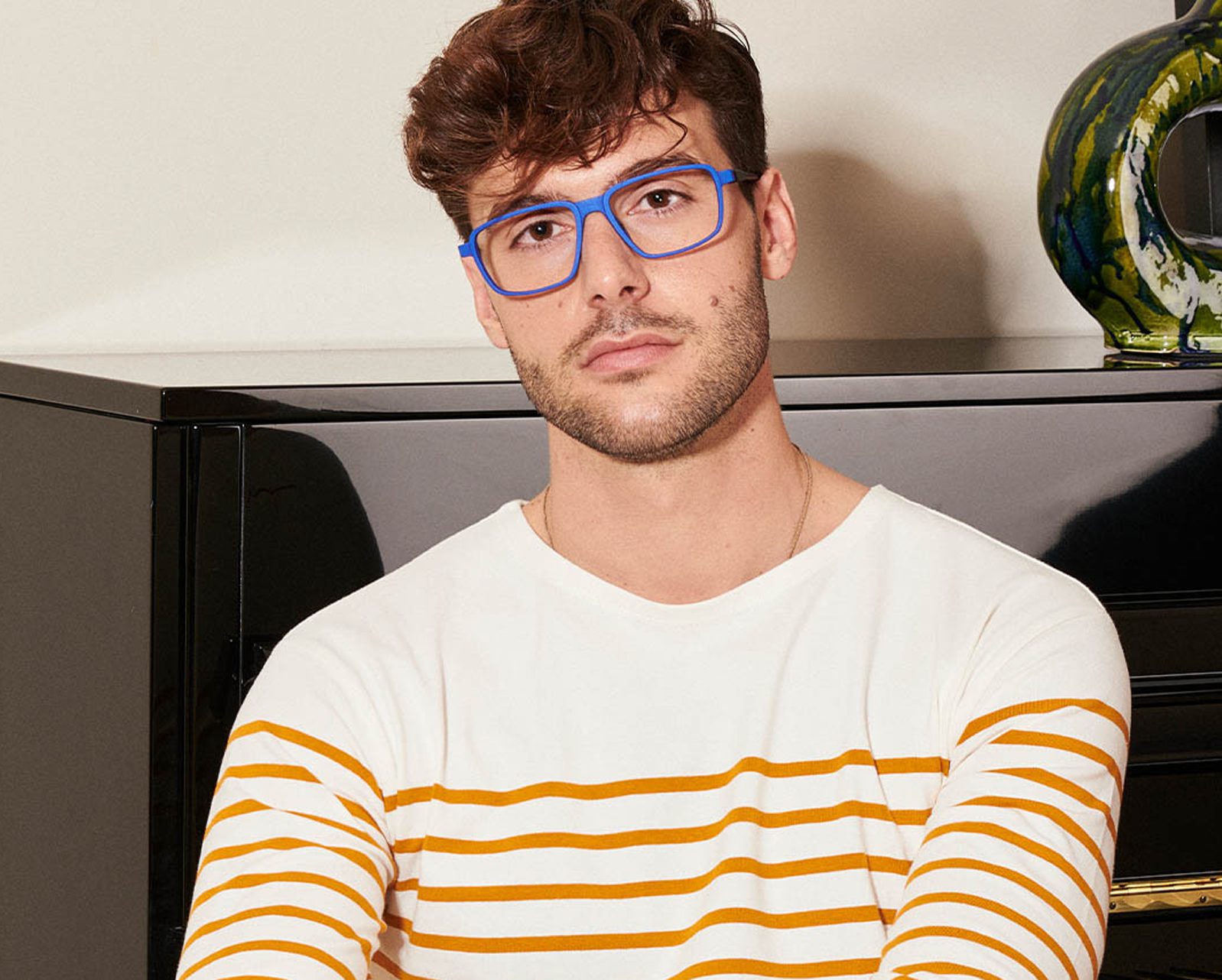 A man with short brown hair and blue glasses from Vu, une lunetterie à Montréal, wears a white shirt with yellow stripes, sitting in front of a black cabinet topped with a colorful ceramic object.